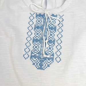 May include: White t-shirt with a blue embroidered design on the front. The design is a geometric pattern with a white string tied at the top.