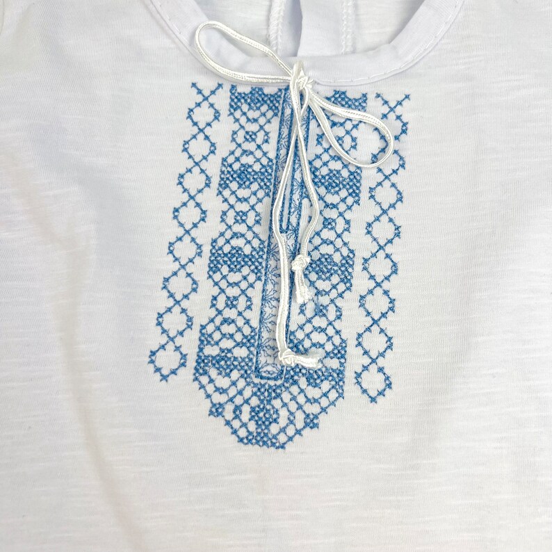 May include: White t-shirt with a blue embroidered design on the front. The design is a geometric pattern with a white string tied at the top.