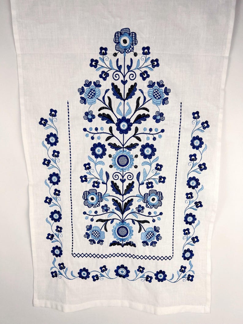 May include: A white rectangular textile featuring intricate embroidery in shades of dark blue, medium blue, and light blue. The design consists of a central vertical floral arrangement flanked by two side borders of small flowers and vines.