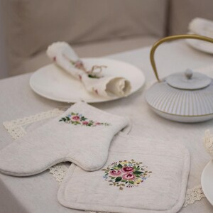 May include: A cream-colored oven mitt and pot holder set with embroidered floral designs in pink, blue, and green. A white teapot with gold accents and a rolled napkin on a plate are also visible.