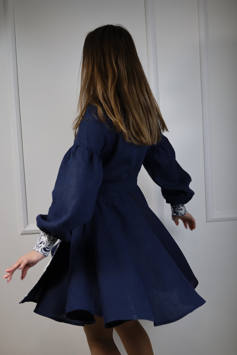 May include: A navy blue dress with long, puffed sleeves and a white and blue patterned trim. The dress has a flared skirt and is made of a soft, flowing fabric.