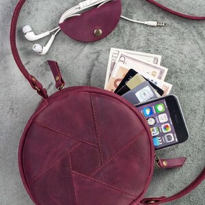 May include: A burgundy leather crossbody bag with a round shape and a geometric design. The bag is open, revealing a smartphone, cash, and credit cards. A matching burgundy leather earbud holder and white earbuds are also present.