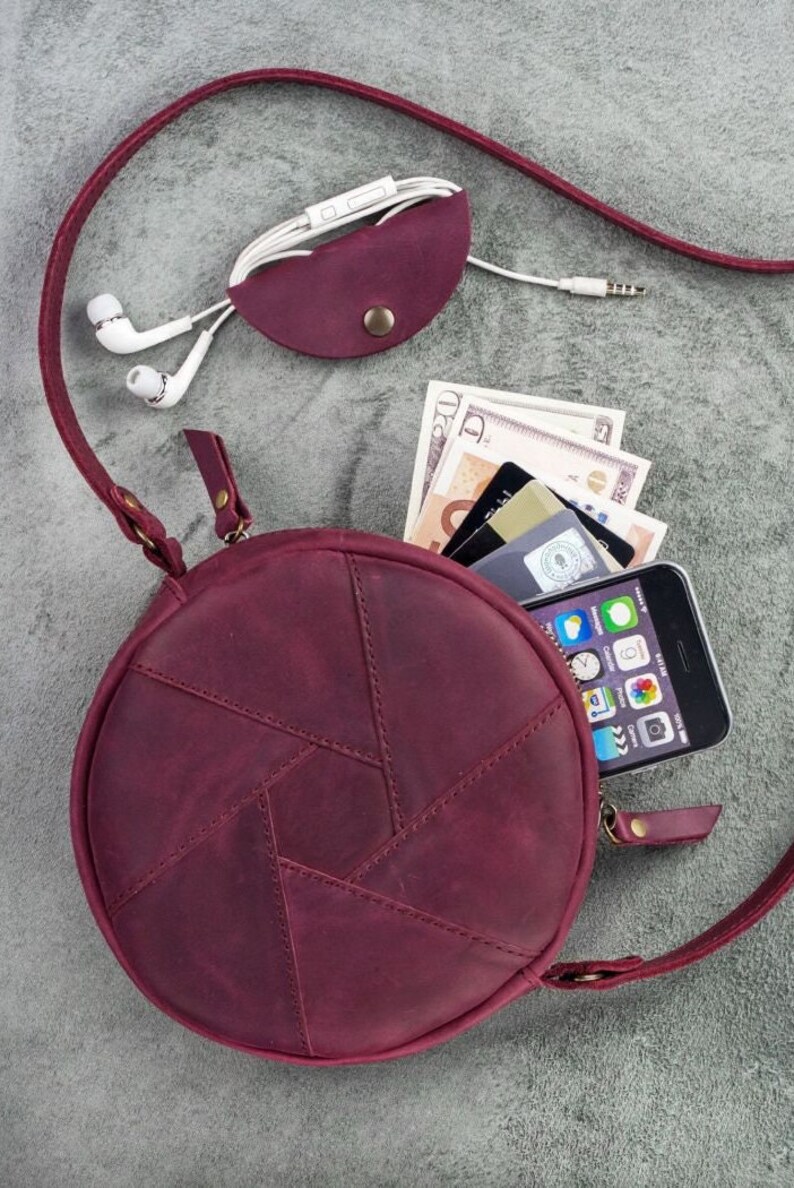 May include: A burgundy leather crossbody bag with a round shape and a geometric design. The bag is open, revealing a smartphone, cash, and credit cards. A matching burgundy leather earbud holder and white earbuds are also present.