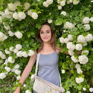 May include: A woman in a gray dress stands in front of a bush of white flowers. She is wearing a beige crossbody bag with a floral embroidery design.