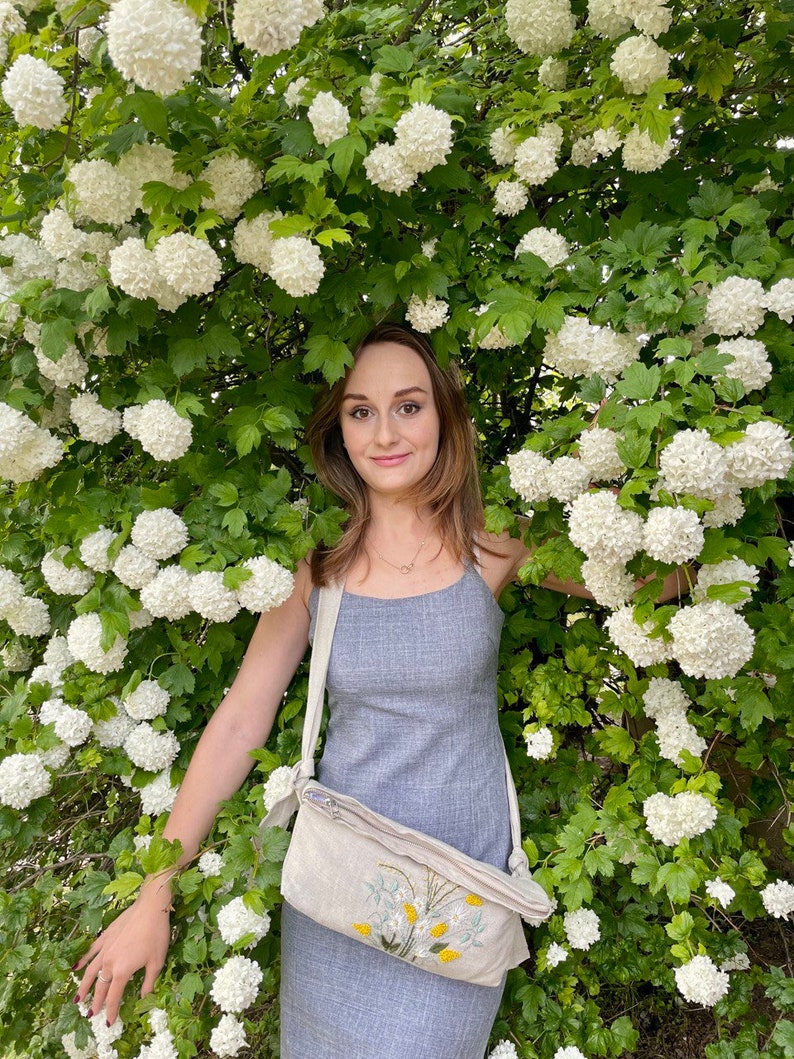 May include: A woman in a gray dress stands in front of a bush of white flowers. She is wearing a beige crossbody bag with a floral embroidery design.