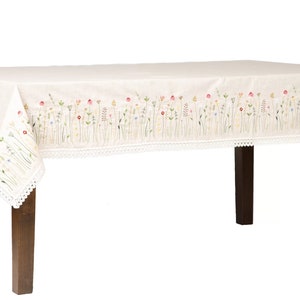 May include: Cream-colored rectangular tablecloth with embroidered floral design in pink, yellow, and green. The edges are trimmed with a white lace border. The tablecloth is draped over a dark brown wooden table.