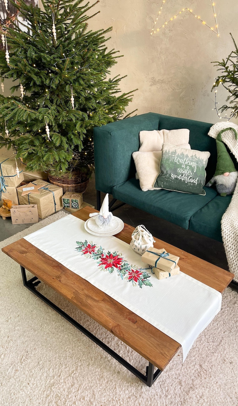May include: A white table runner with a red poinsettia and green holly embroidery design. The runner is on a wooden coffee table with a white fluffy rug underneath. There are wrapped gifts and a small white gnome on the table. A green sofa with cushions is in the background.