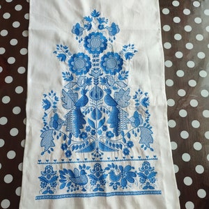 May include: White linen towel with intricate blue floral embroidery. The design features stylized flowers, birds, and foliage, with a delicate crocheted edge. The embroidery is detailed and symmetrical, creating a traditional folk art aesthetic.