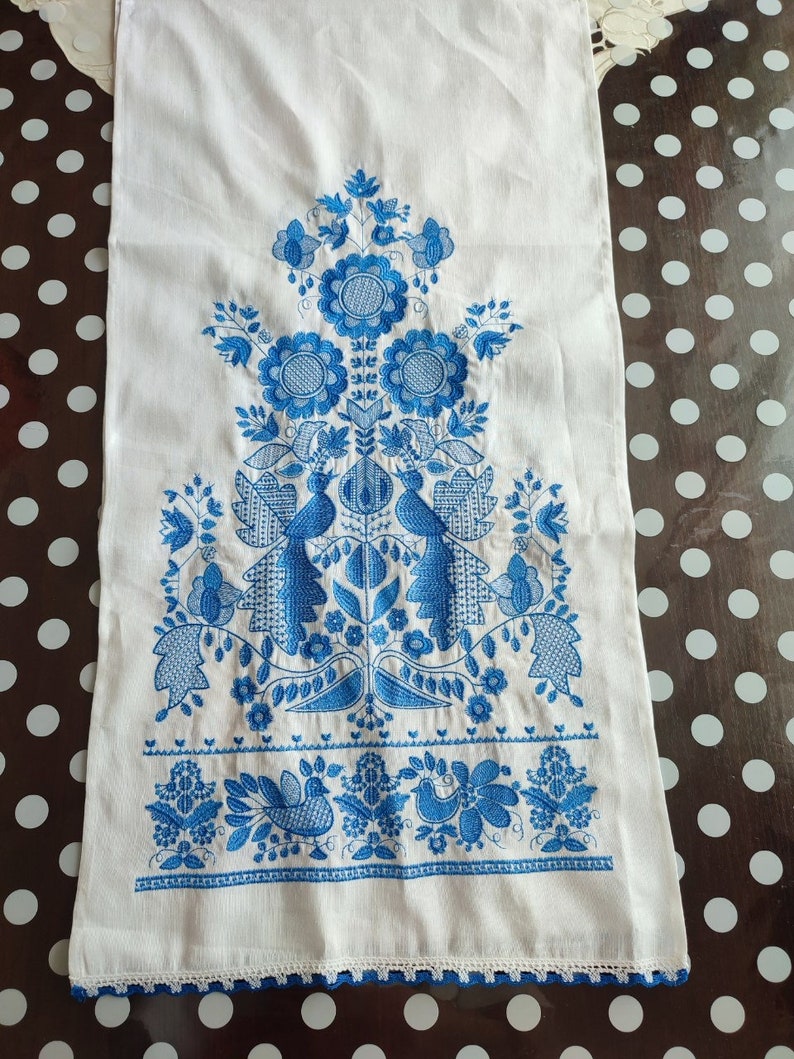 May include: White linen towel with intricate blue floral embroidery. The design features stylized flowers, birds, and foliage, with a delicate crocheted edge. The embroidery is detailed and symmetrical, creating a traditional folk art aesthetic.