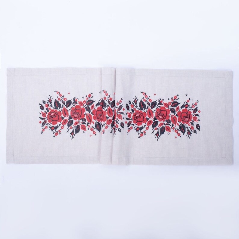 May include: A beige linen table runner with a cross-stitched floral design. The embroidery features red roses and black leaves, creating a symmetrical pattern across the fabric. The runner is rectangular and appears to be folded.