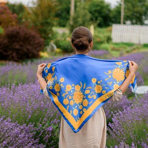 May include: A woman in a field of lavender flowers is wearing a blue and yellow scarf with a sunflower pattern. The scarf has the word "UKRA" printed on it.
