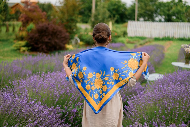 May include: A woman in a field of lavender flowers is wearing a blue and yellow scarf with a sunflower pattern. The scarf has the word "UKRA" printed on it.