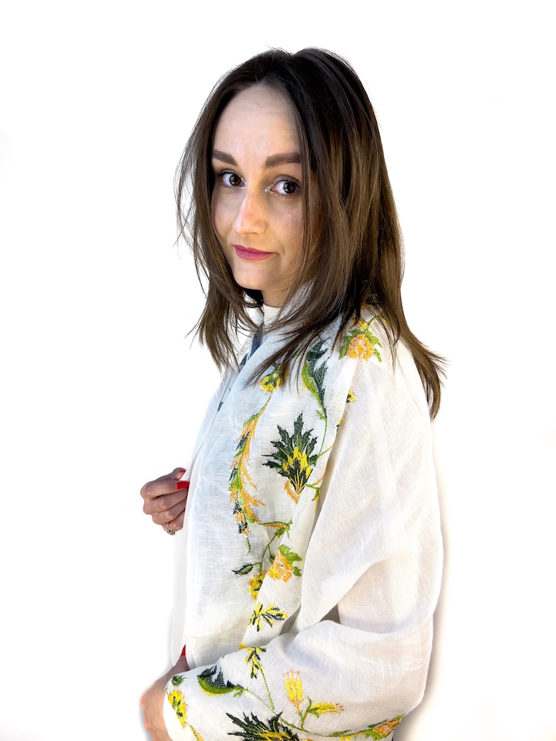 Puede incluir: Una blusa de lino blanca con dise&ntilde;os florales bordados en amarillo y verde. La blusa tiene mangas largas y un corte holgado.
