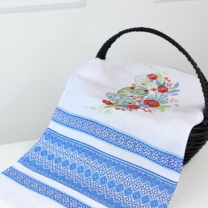 May include: White linen towel with blue geometric embroidery and fringe. The towel features a floral and egg design in the center, placed inside a black woven basket. The towel's border has a detailed blue diamond pattern.