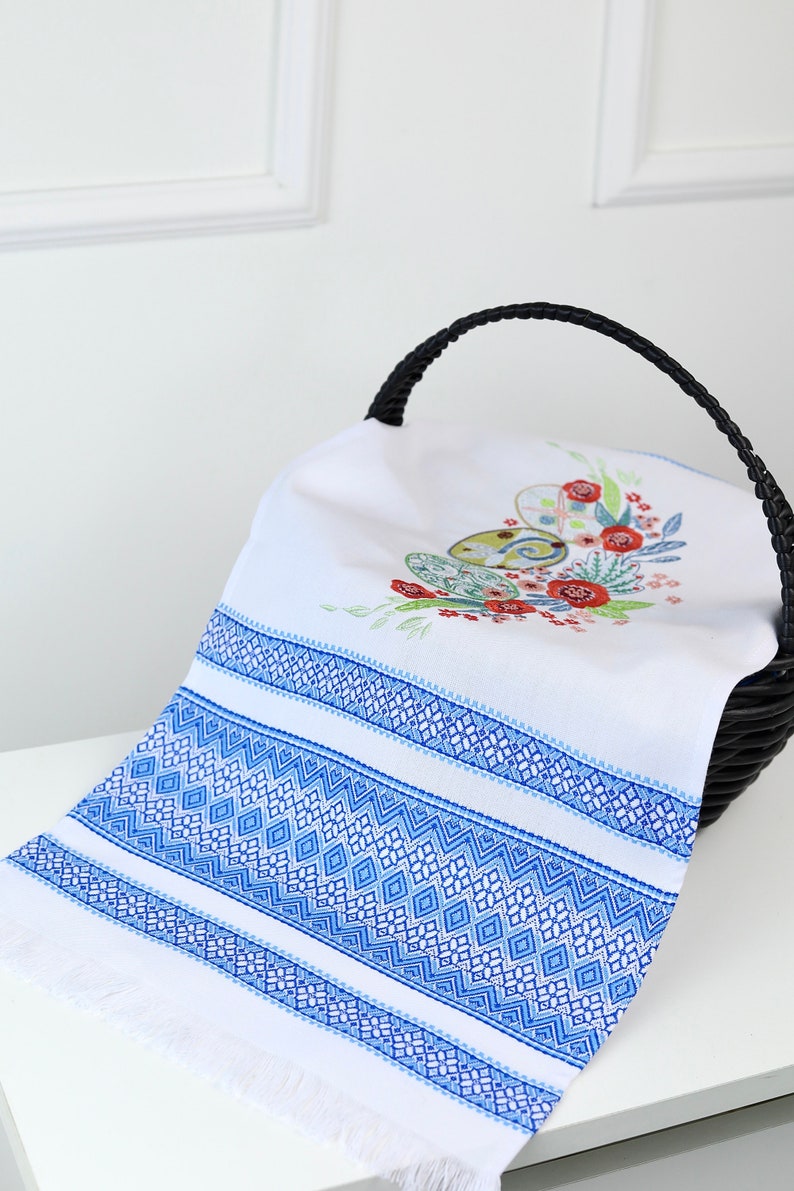 May include: White linen towel with blue geometric embroidery and fringe. The towel features a floral and egg design in the center, placed inside a black woven basket. The towel's border has a detailed blue diamond pattern.