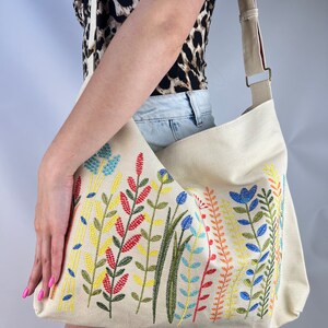 K&ouml;nnte beinhalten: Eine beige Canvas-Tragetasche mit einem langen verstellbaren Riemen. Die Tasche ist mit bunten gestickten Blumen und Bl&auml;ttern verziert.