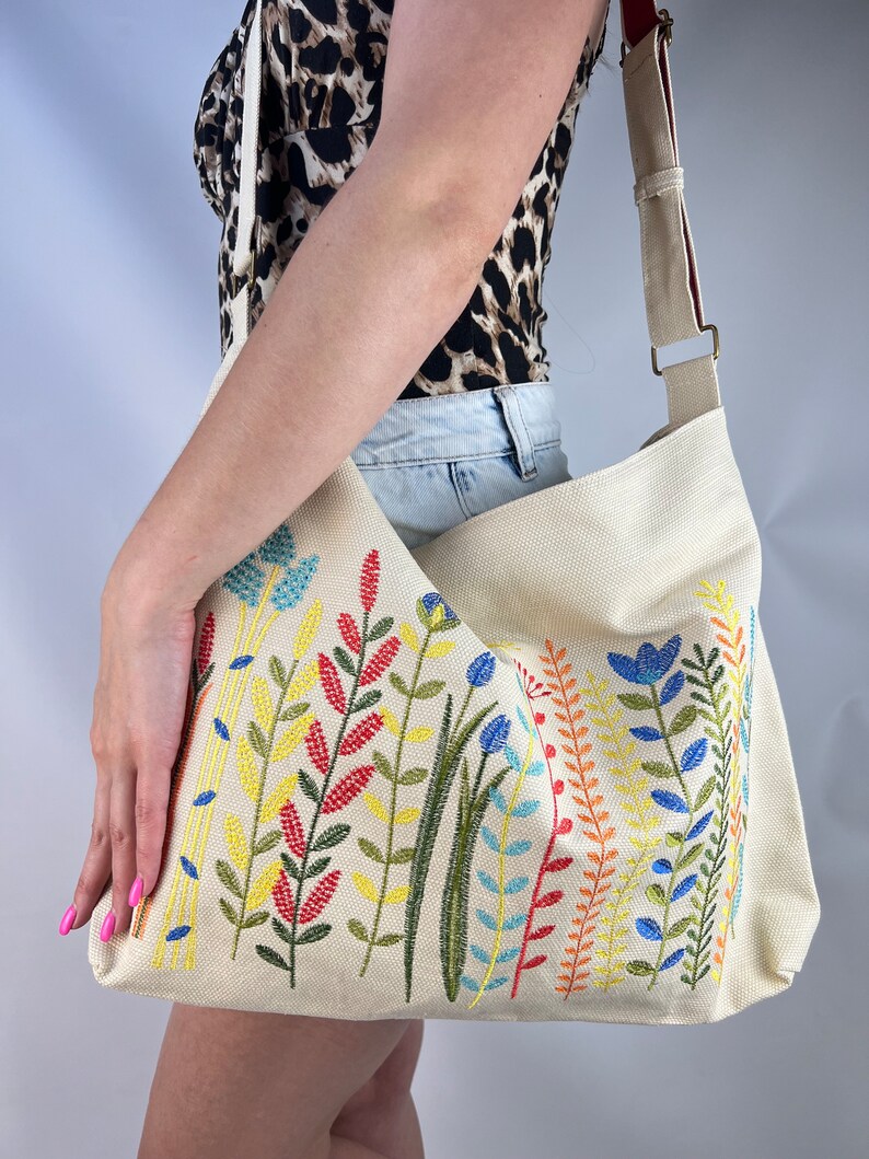 K&ouml;nnte beinhalten: Eine beige Canvas-Tragetasche mit einem langen verstellbaren Riemen. Die Tasche ist mit bunten gestickten Blumen und Bl&auml;ttern verziert.