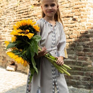 Pu&ograve; includere: Una giovane persona indossa un vestito grigio chiaro con dettagli ricamati e una cintura abbinata, che tiene in mano un grande mazzo di girasoli giallo brillante. L'abito ha maniche lunghe e nappe.