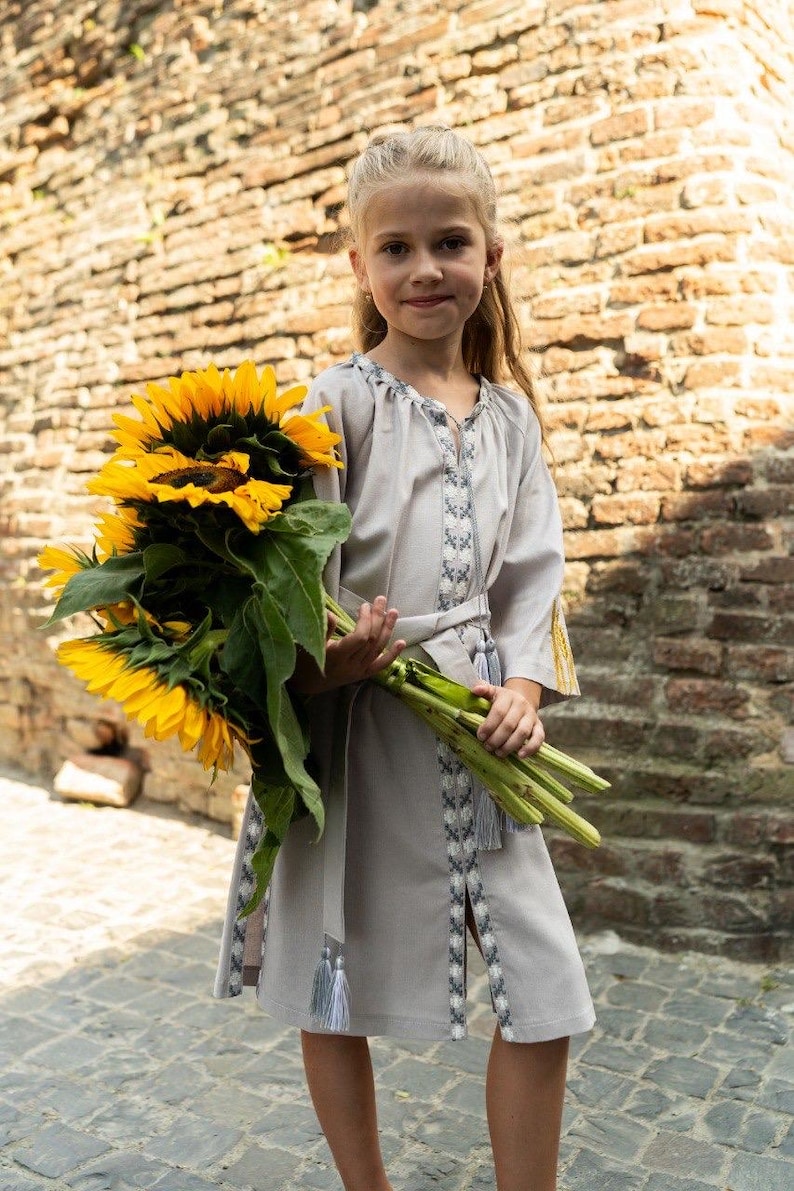 Pu&ograve; includere: Una giovane persona indossa un vestito grigio chiaro con dettagli ricamati e una cintura abbinata, che tiene in mano un grande mazzo di girasoli giallo brillante. L'abito ha maniche lunghe e nappe.