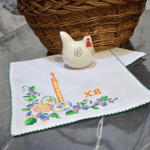 Puede incluir: Una servilleta de lino bordada en blanco con un borde verde. La servilleta presenta una vela, huevos de Pascua y el texto cir&iacute;lico "XB". Una figurita de pollo de cer&aacute;mica blanca est&aacute; encima de la servilleta.