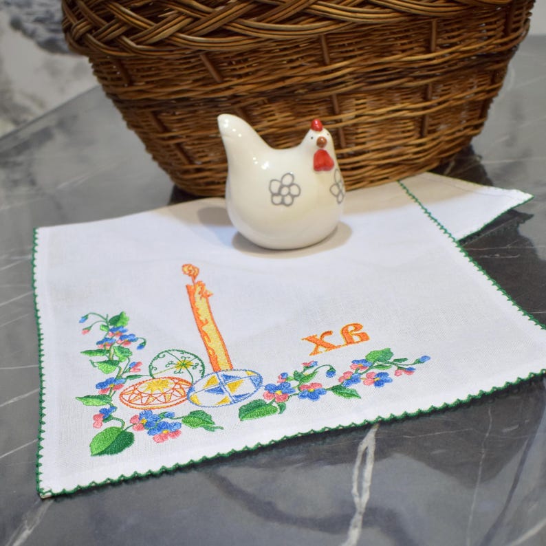 Puede incluir: Una servilleta de lino bordada en blanco con un borde verde. La servilleta presenta una vela, huevos de Pascua y el texto cir&iacute;lico "XB". Una figurita de pollo de cer&aacute;mica blanca est&aacute; encima de la servilleta.