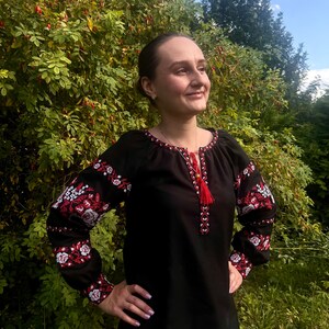 K&ouml;nnte beinhalten: Eine schwarze bestickte Bluse mit roten und wei&szlig;en Blumendesigns. Die Bluse hat lange &Auml;rmel und einen runden Ausschnitt.