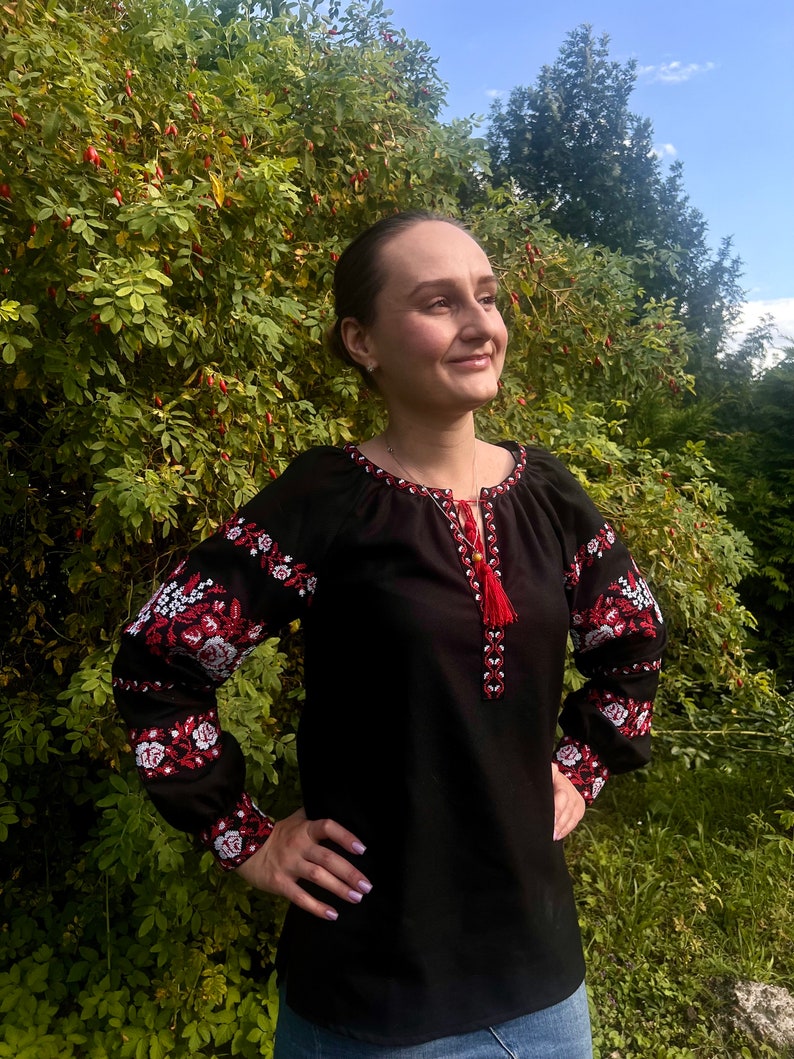 K&ouml;nnte beinhalten: Eine schwarze bestickte Bluse mit roten und wei&szlig;en Blumendesigns. Die Bluse hat lange &Auml;rmel und einen runden Ausschnitt.