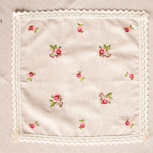 May include: A square, beige linen handkerchief with embroidered pink roses and green leaves. The edges are trimmed with a white lace border. The number "#1" is in the upper left corner.