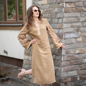 Pode incluir: Vestido de linho bege com detalhes bordados em creme. O vestido apresenta decote em V, mangas compridas com punhos largos e comprimento midi. O vestido &eacute; usado com sand&aacute;lias de salto alto.