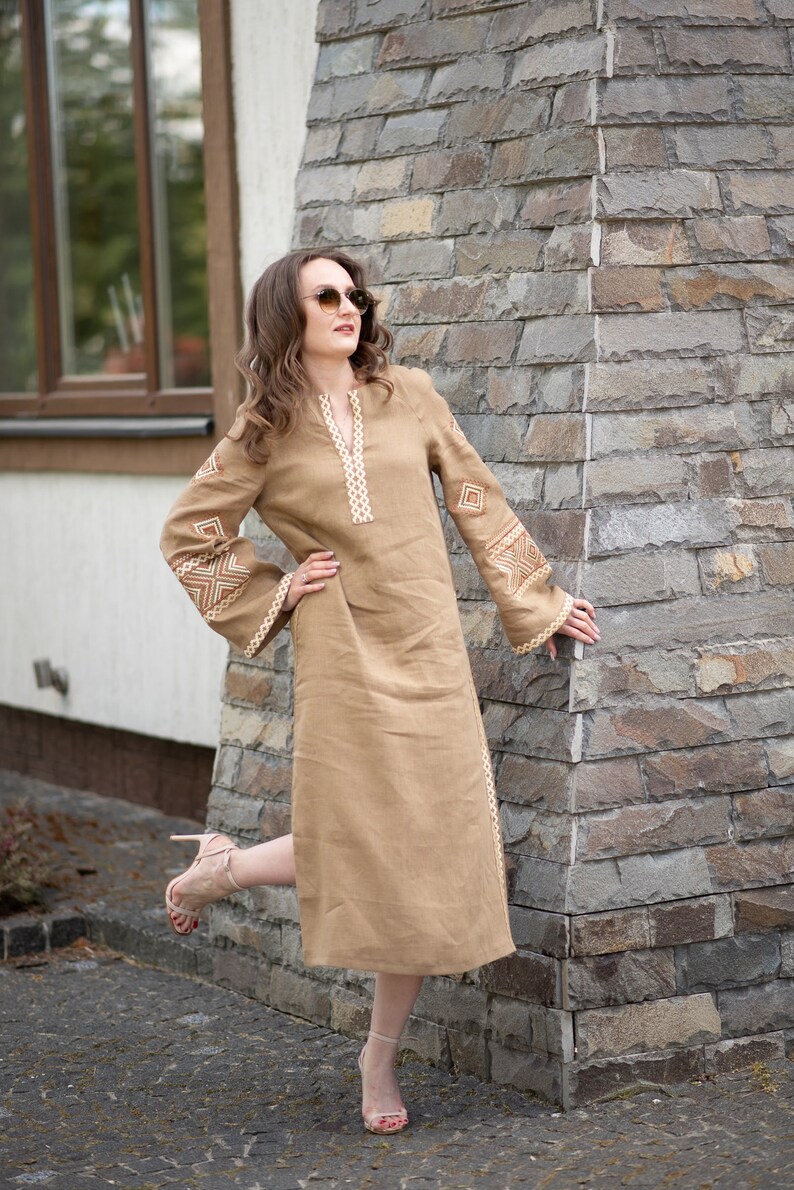 Pode incluir: Vestido de linho bege com detalhes bordados em creme. O vestido apresenta decote em V, mangas compridas com punhos largos e comprimento midi. O vestido &eacute; usado com sand&aacute;lias de salto alto.