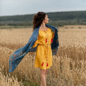 May include: A woman in a yellow dress with red tassels stands in a field of wheat. She is wearing a blue scarf with floral embroidery and is looking off to the side.