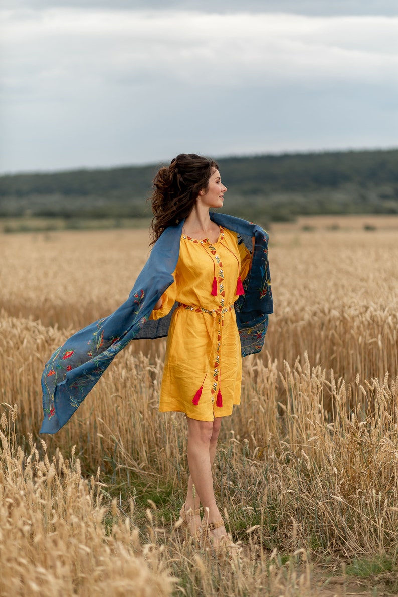 May include: A woman in a yellow dress with red tassels stands in a field of wheat. She is wearing a blue scarf with floral embroidery and is looking off to the side.