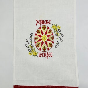 May include: White linen towel with a red lace border. The towel features an embroidered Easter egg design in red, yellow, and green, with Cyrillic text above and below the egg.
