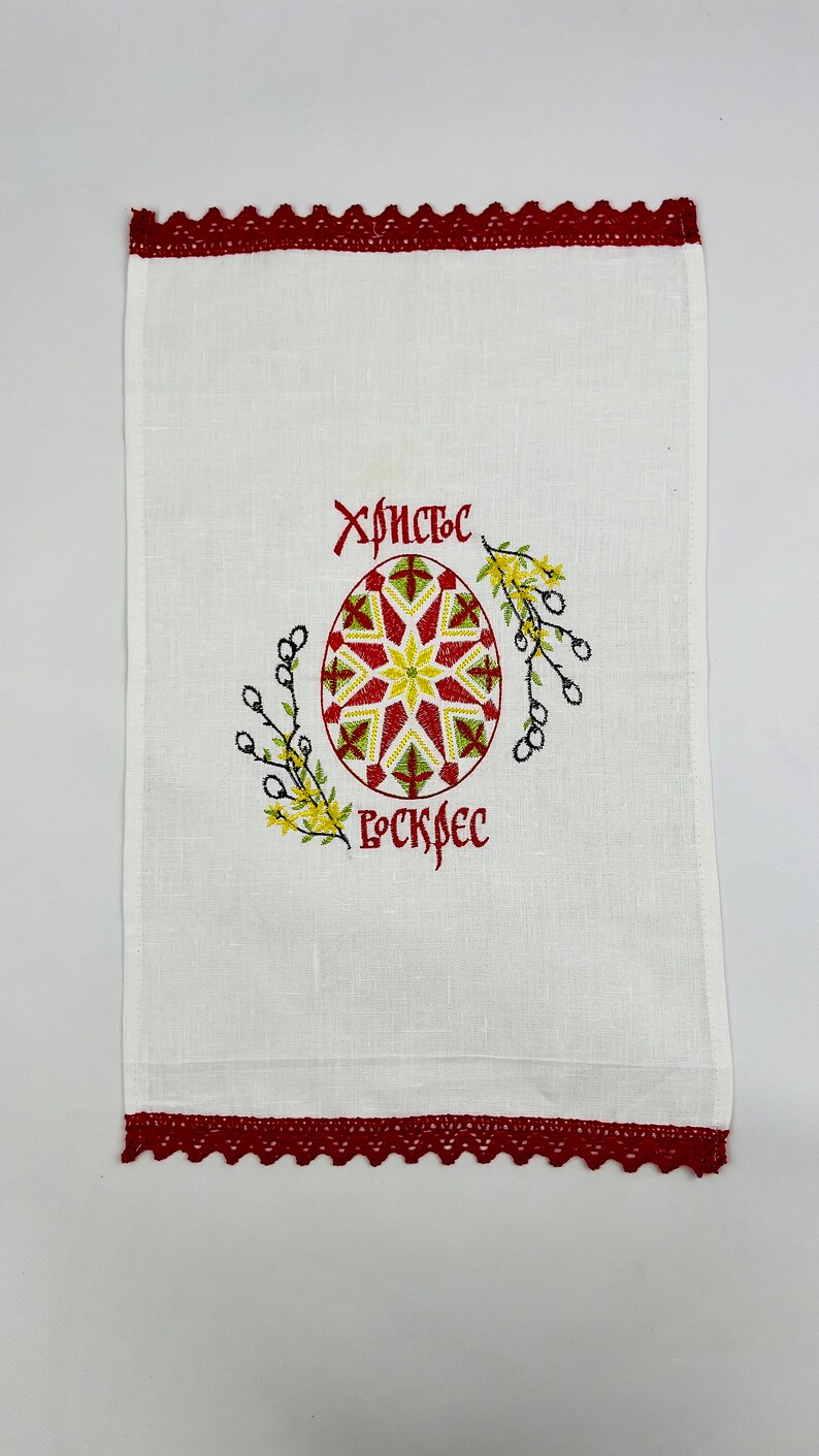 May include: White linen towel with a red lace border. The towel features an embroidered Easter egg design in red, yellow, and green, with Cyrillic text above and below the egg.