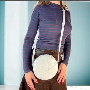 Op de afbeelding: Een witte ronde schoudertas met een geometrisch ontwerp. De tas wordt gedragen door iemand die een lang blauw-rood gestreept shirt en een bruine rok draagt.