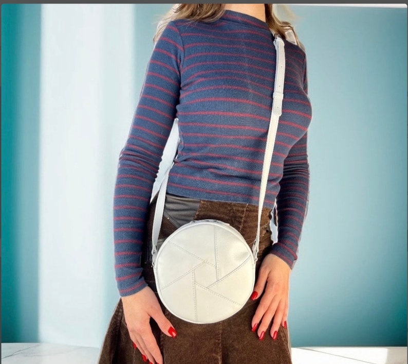 Op de afbeelding: Een witte ronde schoudertas met een geometrisch ontwerp. De tas wordt gedragen door iemand die een lang blauw-rood gestreept shirt en een bruine rok draagt.