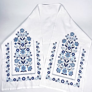 May include: A white linen table runner with blue and white floral embroidery. The design features two vertical floral motifs with a border of smaller flowers.