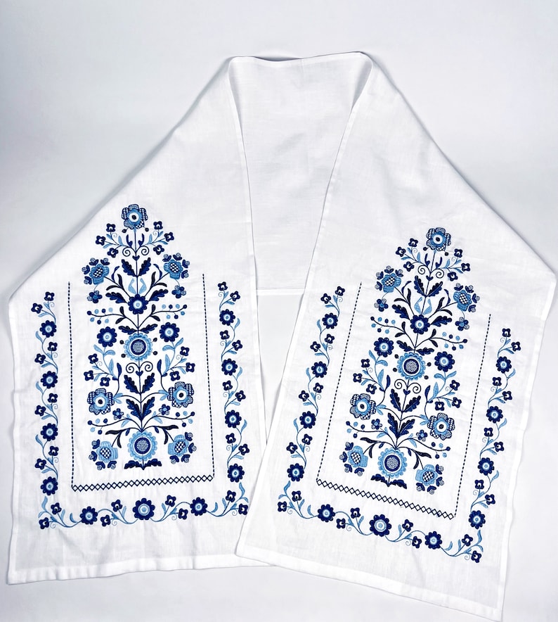 May include: A white linen table runner with blue and white floral embroidery. The design features two vertical floral motifs with a border of smaller flowers.