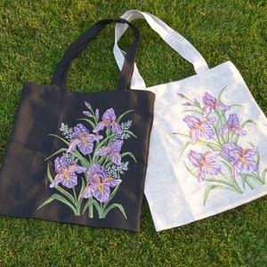 Op de afbeelding: Twee tote bags met geborduurde irisbloemen. E&eacute;n tas is zwart met paarse irissen en de andere is wit met paarse irissen.
