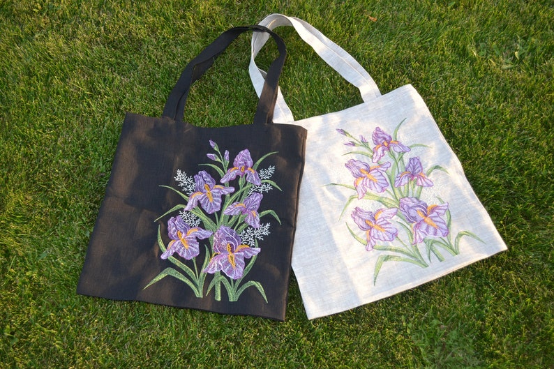 Op de afbeelding: Twee tote bags met geborduurde irisbloemen. E&eacute;n tas is zwart met paarse irissen en de andere is wit met paarse irissen.