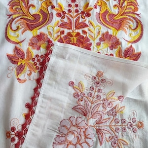 Peut inclure: Textile blanc brod&eacute; avec un motif vibrant. La broderie pr&eacute;sente deux oiseaux stylis&eacute;s dans des tons de jaune, rouge et orange, ainsi que des motifs floraux et de baies. Un bord rouge festonn&eacute; borde le textile, ajoutant une touche d&eacute;corative.