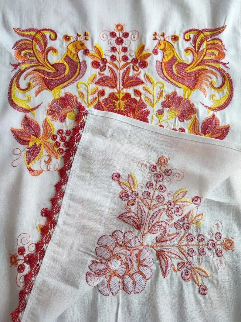 Peut inclure: Textile blanc brod&eacute; avec un motif vibrant. La broderie pr&eacute;sente deux oiseaux stylis&eacute;s dans des tons de jaune, rouge et orange, ainsi que des motifs floraux et de baies. Un bord rouge festonn&eacute; borde le textile, ajoutant une touche d&eacute;corative.