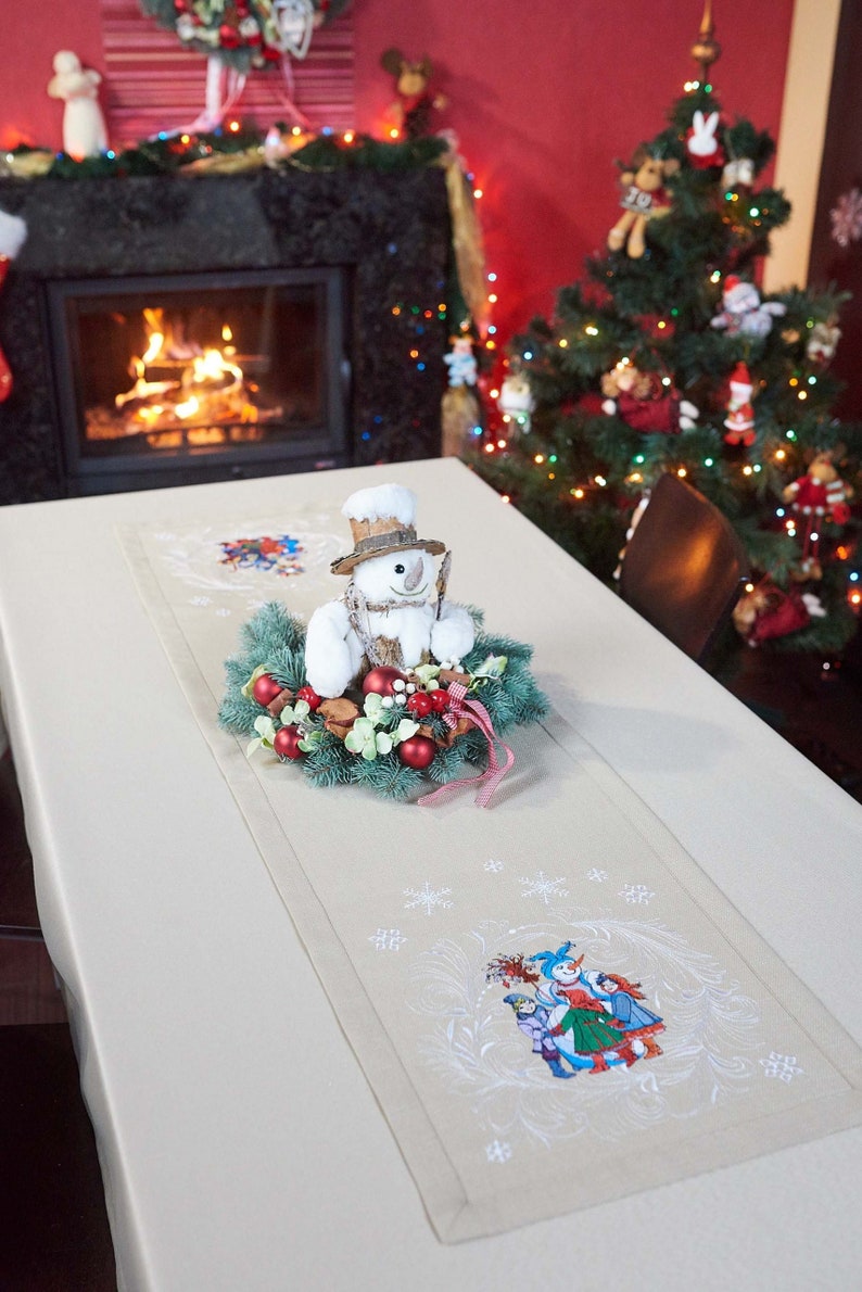 Op de afbeelding: Beige kersttafelloper met geborduurde sneeuwvlokken en een winters tafereel. Een sneeuwmanversiering staat op een krans van groen en rode bessen. Een versierde kerstboom en open haard zijn op de achtergrond.