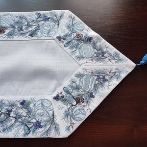 以下が含まれることがあります： 松の枝、ベリー、オーナメントが冬のホリデーをテーマにした青い刺繍の縁取りが施された白いテーブルランナー。ランナーには青いタッセルが付いています。