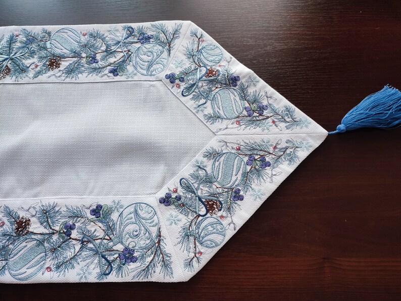 以下が含まれることがあります： 松の枝、ベリー、オーナメントが冬のホリデーをテーマにした青い刺繍の縁取りが施された白いテーブルランナー。ランナーには青いタッセルが付いています。