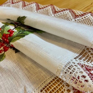 May include: White linen table runner with red and green embroidered holly berries and leaves. The runner has a white lace border with red accents.