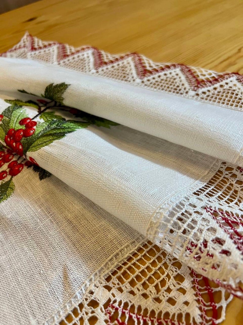May include: White linen table runner with red and green embroidered holly berries and leaves. The runner has a white lace border with red accents.