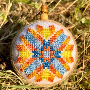 May include: A round, white, fabric ornament with a colorful geometric pattern in shades of orange, yellow, blue, and white. The ornament is stitched with a cross-stitch pattern and has a wooden bead at the top.