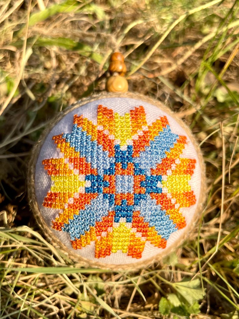 May include: A round, white, fabric ornament with a colorful geometric pattern in shades of orange, yellow, blue, and white. The ornament is stitched with a cross-stitch pattern and has a wooden bead at the top.