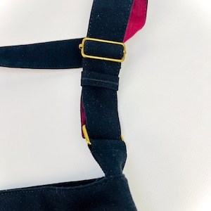 K&ouml;nnte beinhalten: Eine schwarze Canvas-Tragetasche mit einem schwarzen und roten Riemen und einer goldenen Schnalle. Der Riemen ist verstellbar.